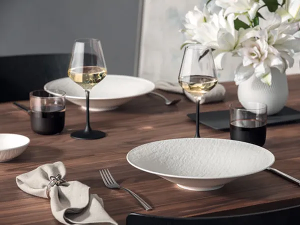 Manufacture Esstisch mit Tellern, Gläsern und Vase Ein mit weißen Manufacture-Tellern, Weingläsern, Besteck und einer Serviette gedeckter Esstisch. Im Hintergrund steht eine Vase mit weißen Blumen von Manufacture.