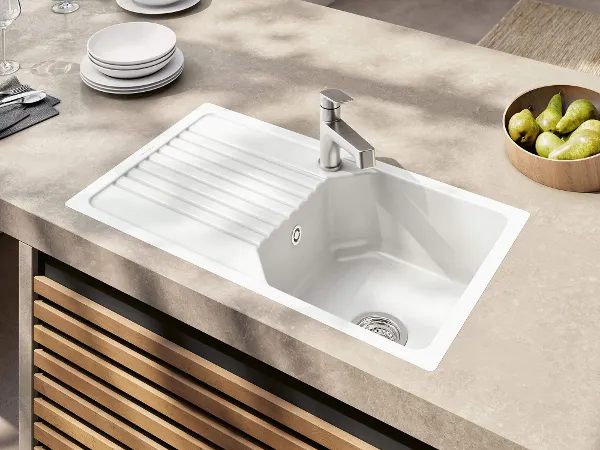 White Linara kitchen sink with beige worktop A white kitchen sink with an elegant tap is installed in a stone worktop, next to a bowl of pears and stacked plates.