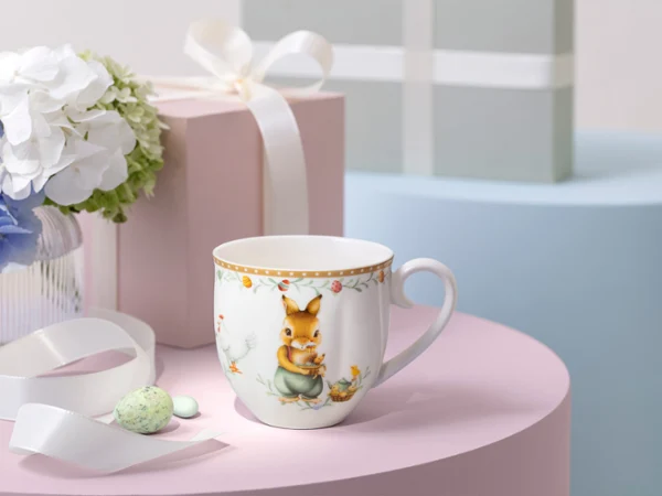 Une tasse décorée Annual Easter Edition posée sur une surface rose à côté d’un ruban blanc, de boîtes-cadeaux pastel et d’un vase de fleurs.