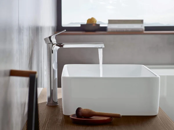 Une robinetterie moderne en chrome dirige l’eau vers un lavabo rectangulaire blanc posé sur une tablette en bois dans une salle de bains minimaliste avec une fenêtre en arrière-plan.