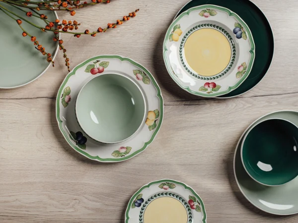 French Garden with Perlemor Alga An overhead view of mixed dinnerware, featuring solid green bowls and plates alongside white plates with a colorful fruit border, arranged on a light wooden surface