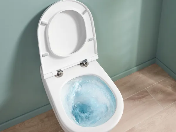 Universo TwistFlush in funzione, con una parete di colore verde chiaro WC bianco con copriwater sollevato con acqua di risciacquo blu, di fronte a una parete blu-verde e a un pavimento in legno chiaro.