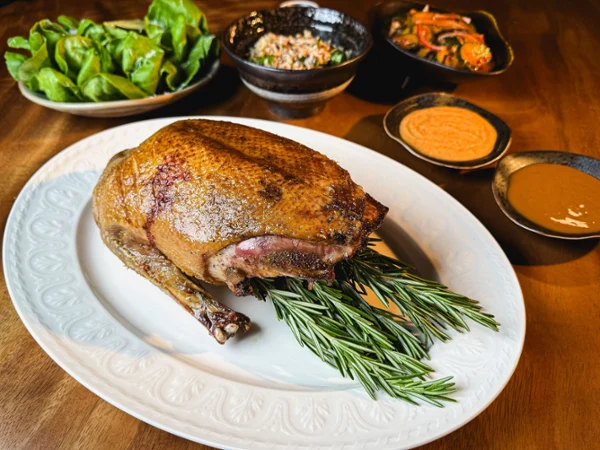 Golden-brown roasted duck rests on a white platter with rosemary, surrounded by greens, kimchi, rice, and dipping sauces on a warm wood table.