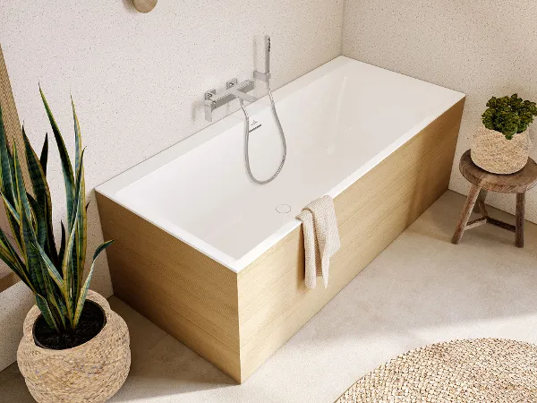 Modernes Badezimmer mit einer rechteckigen Badewanne aus der Kollektion Subway von Villeroy & Boch Ein modernes Badezimmer mit einer rechteckigen Badewanne aus der Kollektion Subway von Villeroy & Boch, einem Holzhocker mit einer Pflanze, einem gewebten Teppich und einer großen Topfpflanze neben der Wanne. Die Wände haben ein hell gesprenkeltes Finish mit runden Holzakzenten.