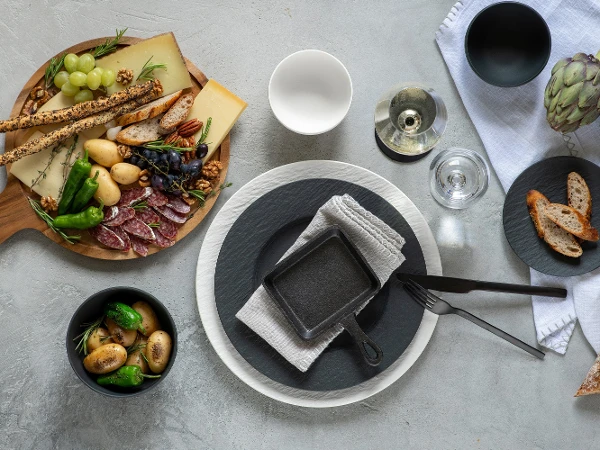 Artesano and Manufacture plates at the raclette A “Manufacture” place setting from Villeroy & Boch with plate, napkin, fork, knife, and glass is beautifully arranged. Next to it is a charcuterie board adorned with cheese, bread, meat, grapes, and olives. A small bowl of potatoes joins other elegantly designed bowls in the display.