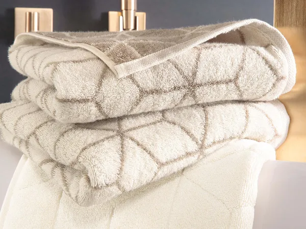 Un aménagement de salle de bains élégant avec des serviettes « Carré » beiges et un robinet doré. Une pile de serviettes « Carré » beiges pliées à motifs géométriques de Villeroy & Boch, placées à côté d’un robinet doré.