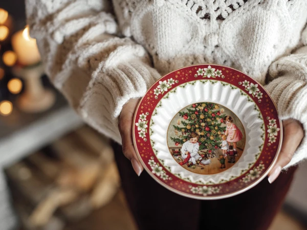 A person wearing a cozy sweater holds a decorative plate with a Christmas scene of children near a tree. Warm, festive ambiance enhances holiday spirit.