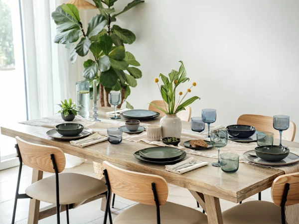 Dining table set with varios items from the Lave collection by Villeroy & Boch The dining table set with varios items from the Lave collection by Villeroy & Boch in green and grey, accentuated by blue glasses and napkins. A vase of yellow flowers and a small potted plant provide decoration, while a large plant stands by the window.