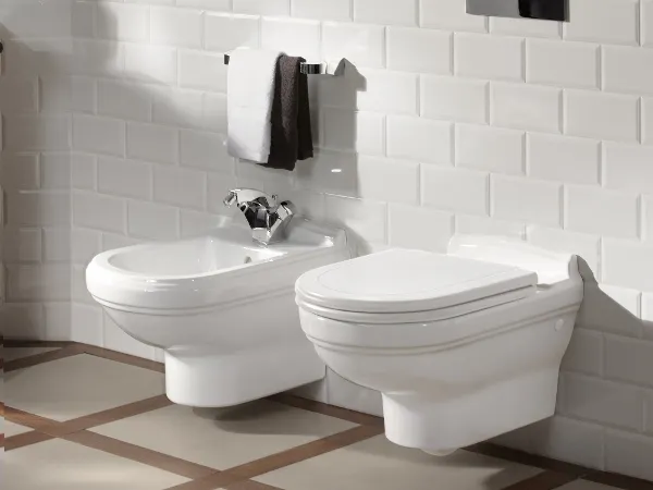 Gefliestes Badezimmer mit weißer Toilette und Bidet der Kollektion Hommage Ein Badezimmer mit weiß gefliesten Wänden und weiß gefliestem Boden, in dem nebeneinander ein Bidet und eine Toilette installiert sind. Darüber hängen drei gerahmte Silhouettenportraits an der Wand. Über dem Bidet hängt ein Handtuch.