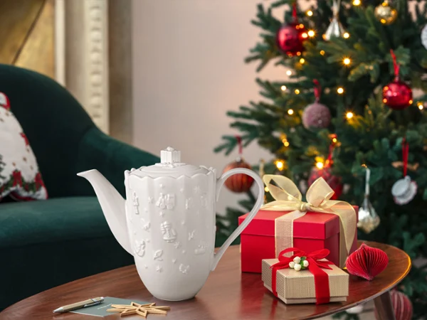 Two wrapped gifts and a white Toys Delight Royal Classic ceramic jug stand on a wooden table next to a decorated Christmas tree.