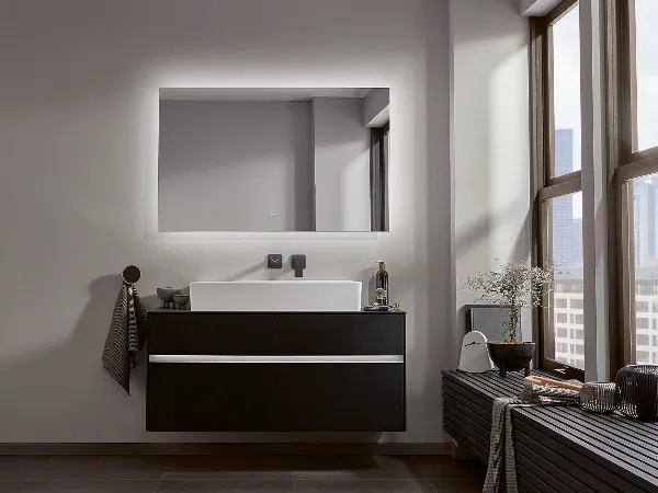 Modernes Badezimmer mit schwebendem schwarzem Collaro Waschtisch von Villeroy & Boch Modernes Badezimmer mit schwebendem schwarzem Collaro Waschtisch von Villeroy & Boch, beleuchtetem Spiegel, Handtuch an einem runden Haken, Topfpflanzen und einer Bank am Fenster.