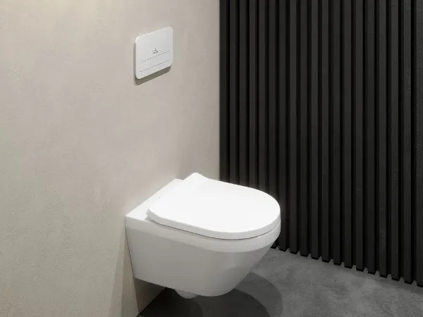 White Architectura toilet with a rectangular white flush button A modern wall-mounted white Architectura toilet with a rectangular flush button above, in front of a beige wall with black slatted panelling. The floor is made of grey concrete.