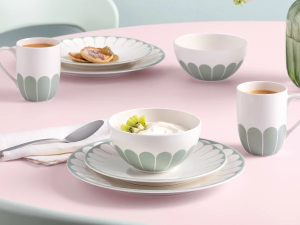 A table set with plates, bowls, and mugs featuring a flower pattern, with food and a Fleur glass vase on a pink tablecloth.