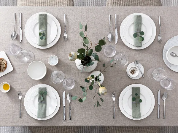 Modern dining table for six people with white Manufacture plates Modern dining table for six people with white Manufacture plates, cutlery and green floral decoration, seen from above.