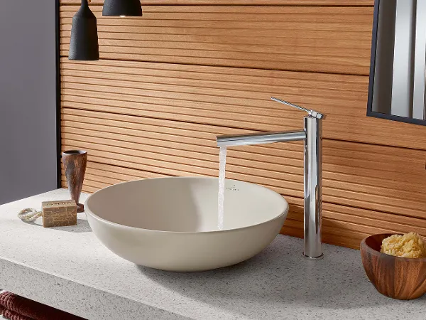 Une robinetterie Loop & Friends avec un lavabo rond dans une salle de bains moderne Une salle de bains moderne avec une vasque à poser ronde Loop & Friends, de l’eau s’écoule d’un robinet minimaliste, des panneaux muraux en bois et divers articles de toilette sur un plan de toilette gris.
