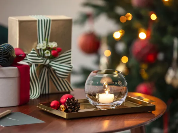 A burning candle in a Toy's Delight tea light holder A burning candle in a Toy's Delight tea light holder stands on a tray next to wrapped presents, with a decorated Christmas tree in the background.