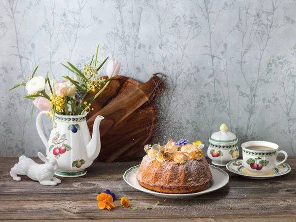 French Garden coffee and tea with Artesano platter A decorated Bundt cake, a cup of tea, a sugar bowl, a floral teapot with fresh flowers, and a ceramic rabbit figurine are arranged on a wooden table against a floral wallpaper.