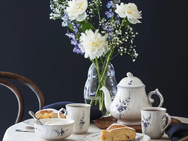 Old Luxembourg porcelain on a coffee table A table covered with a white tablecloth for afternoon tea, with an Old Luxembourg floral-patterned teapot, cups, plates with slices of cake and a vase of white and purple flowers against a dark background.