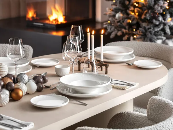 Festlicher Esstisch mit weißem Afina-Geschirr Ein festlicher Esstisch mit weißem Afina-Geschirr, Kerzen und dekorativen Ornamenten von Villeroy & Boch, neben einem brennenden Kamin und einem geschmückten Weihnachtsbaum im Hintergrund.