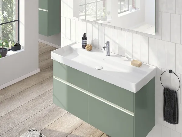 Un lavabo Collaro blanc avec un plan de toilette vert et grand miroir Une salle de bains moderne avec un grand miroir, un plan de toilette vert avec un lavabo blanc, du savon et un flacon sur la tablette. Une serviette est suspendue à un anneau porte-serviette noir. La lumière naturelle passe à travers une fenêtre.