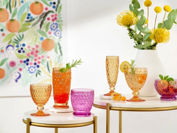 Bunte Boston Gläser auf Tischen mit verschiedenen Getränken Bunte Gläser auf Tischen mit verschiedenen Getränken, garniert mit Kräutern und Fruchtscheiben. Gelbe Blumen und Grünpflanzen in einer Vase. Wandkunst mit Blumen- und Fruchtmustern im Hintergrund.