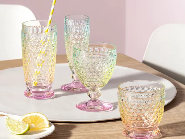 Vier Boston Pearl-Gläser mit Zitronenscheiben Vier Gläser aus strukturiertem Glas von Villeroy & Boch Boston Pearl, drei davon leer und eines mit einem gelb gestreiften Strohhalm, stehen auf einem weißen Tablett auf einem Holztisch. Daneben steht ein kleiner Teller mit Zitronenscheiben.