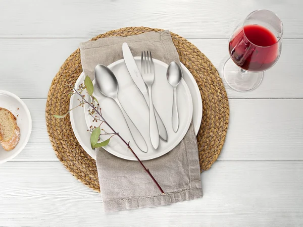 Arthur Besteck auf einem weißen Artesano Teller von Villeroy & Boch Ein gedeckter Tisch mit einem Artesano Teller, Arthur Besteck und einer Serviette auf einem runden Tischset. Daneben stehen ein Glas Rotwein und ein kleiner Teller mit Brotscheiben. Auf dem Teller liegt ein Zweig.