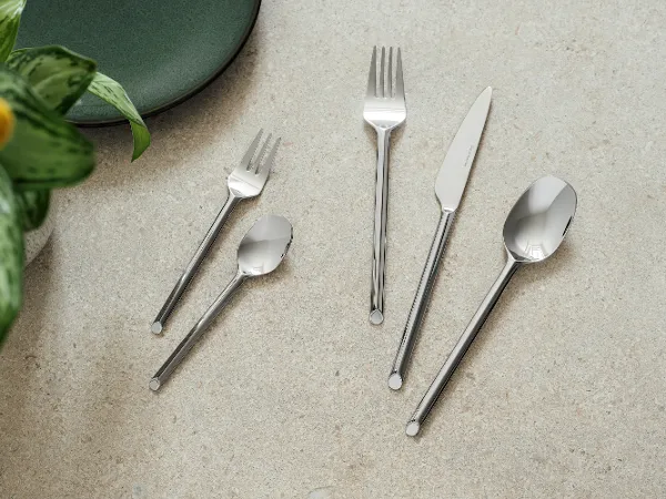 Cinq couverts like de like.by Villeroy & Boch- fourchette, couteau, cuillère, fourchette à dessert et cuillère à café Cinq couverts like. Like. by Villeroy & Boch - fourchette, couteau, cuillère, fourchette à dessert et cuillère à café - sont disposés sur une surface en pierre claire à côté d'une assiette verte et d'une plante.