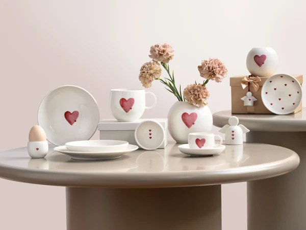 Stoviglie in ceramica di With Love con motivi a cuore rosso, disposte su tavoli rotondi con fiori e una confezione regalo.