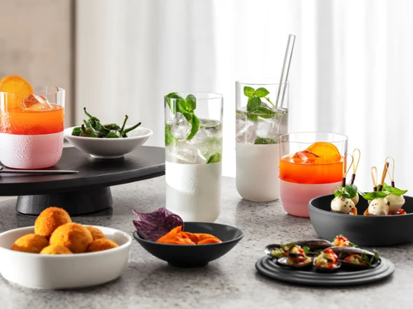 Assorted appetizers and refreshing cocktails served in modern frosted glassware and minimalist bowls on a stone countertop. The spread features citrus cocktails, sparkling mint drinks, savory bites, and colorful snacks, creating a stylish setting for contemporary entertaining.
