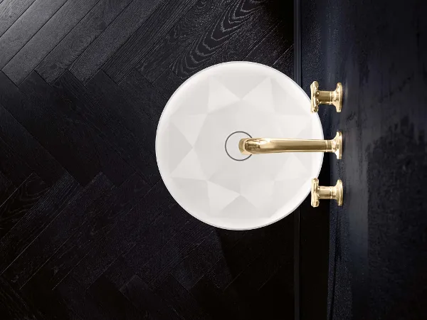 Octagon wastafel met gouden kraan Bovenaanzicht van een witte, gefacetteerde wastafel met gouden kraan en handgrepen, gemonteerd aan een zwarte muur. De vloer is gemaakt van donker visgraathout.