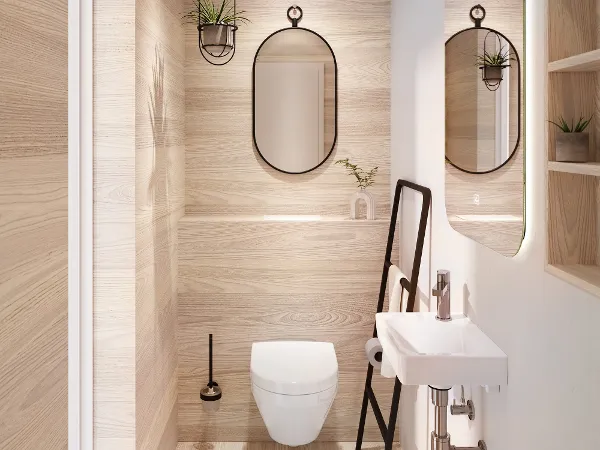 Villeroy & Boch Architectura toilet in white A modern bathroom with light-coloured wood panelling, a wall-mounted toilet, a round mirror, a small washbasin with exposed pipes and a black towel ladder. There are shelves with plants and toiletries.