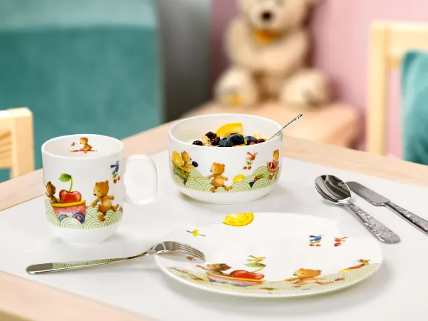 De la vaisselle pour enfants Happy as a bear avec des fruits et des biscuits Un ensemble pour le petit-déjeuner Hungry as a bear avec un bol de céréales et des fruits sur une table, ainsi qu’une tasse, des couverts de table et un ours en peluche en arrière-plan.