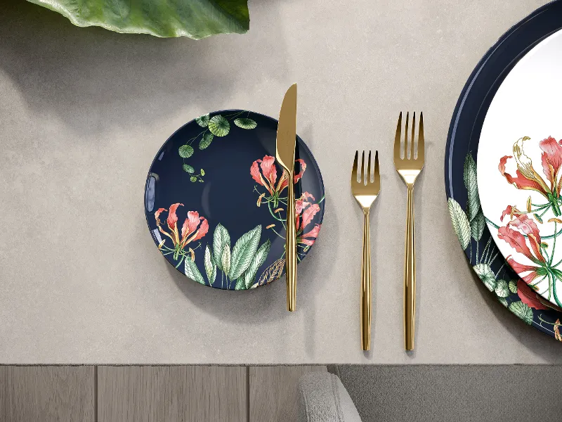 Ein Avarua-Teller von Villeroy & Boch mit goldenem Besteck steht neben einem größeren, passenden Blumenteller auf einem grauen Tisch.