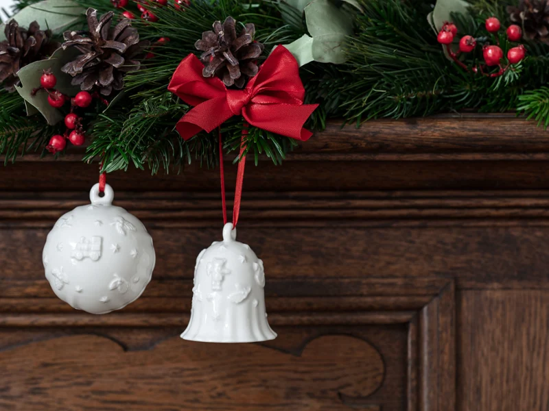 The white Toy’s Delight Royal Classic ball and bell ornaments from Villeroy & Boch hang from red ribbons on a garland decorated with pine cones and red berries, which is draped over a wooden surface.