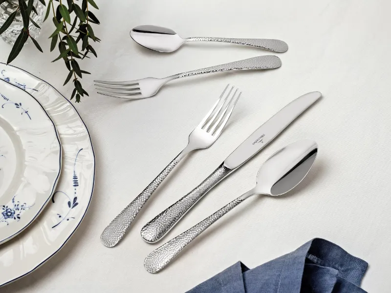 A Leandra flatware set from Villeroy & Boch is elegantly arranged on a white table. It consists of two forks, two spoons, and a knife. Plates with a blue rim and a blue napkin are partially visible, while green foliage lends the table a decorative charm.