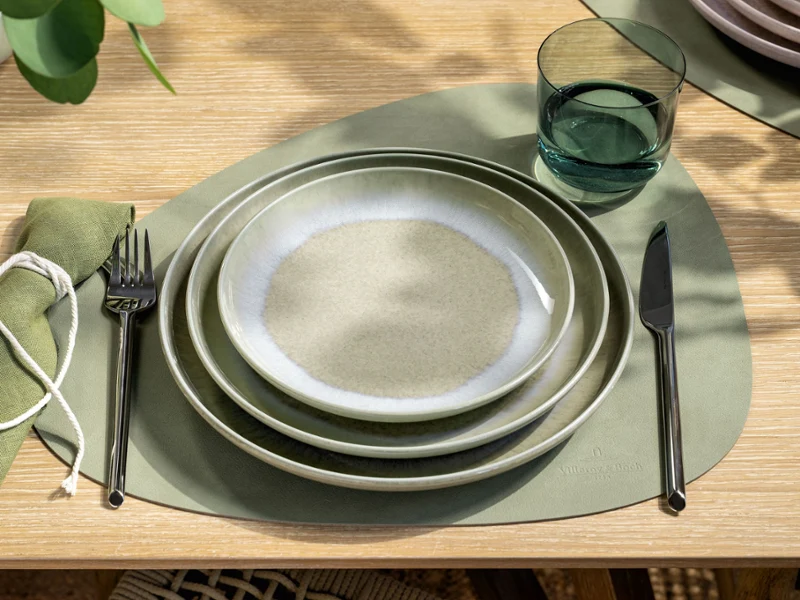 Top-down view of a table setting with Villeroy & Boch’s Perlemor Alga plates in layered shades of soft green, combined with a Like Sage water glass. The setting reflects a naturally calm, earthy aesthetic with handcrafted character and subtle tonal variations.