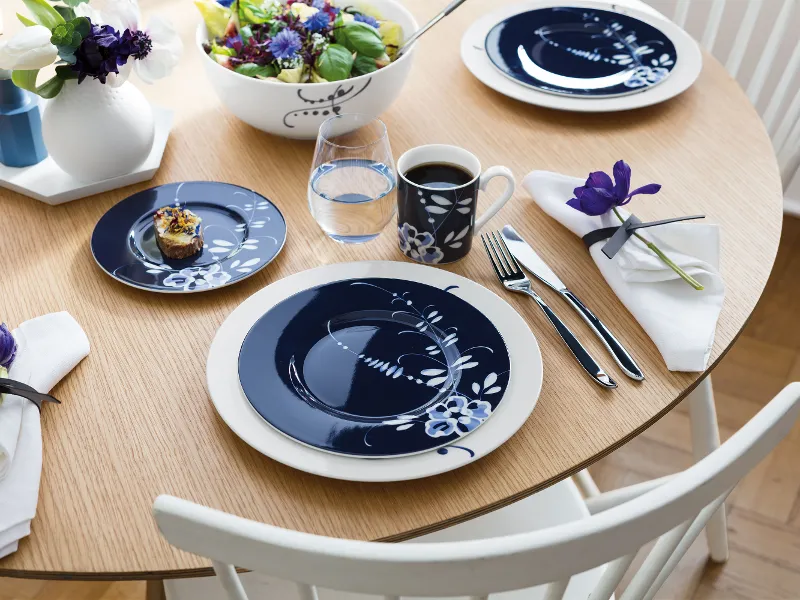 Een ronde houten tafel met blauwe en witte borden, kopjes en een klein dessert. In het midden staat een slakom en blauwe bloemen.