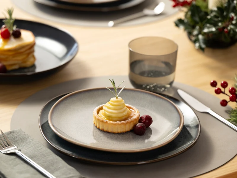 A dessert tart garnished with cream and berries sits on a Lave gray plate, next to a glass of water and utensils.