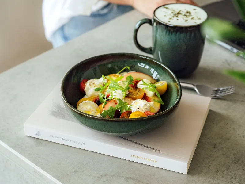 Un'insalata con pomodori, lattuga e formaggio è presentata su un libro in una ciotola verde Lave di Villeroy & Boch, accompagnata da una bevanda schiumosa in una tazza verde abbinata su sfondo grigio.