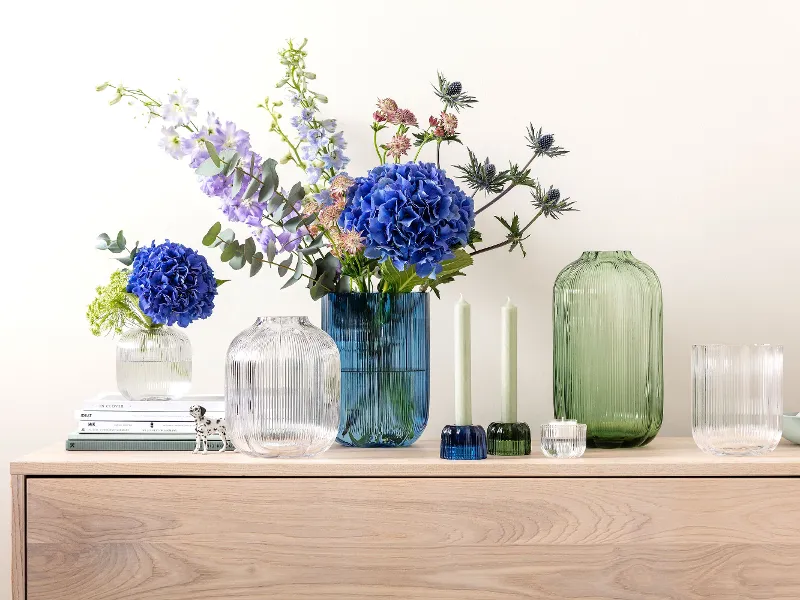 Ein Sideboard mit Fleur-Vasen von Villeroy & Boch, geschmückt mit blauen und violetten Blumen, neben zwei grünen Kerzen und gestapelten Büchern.