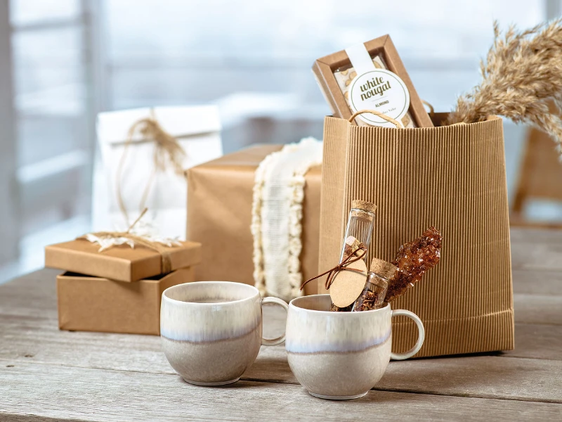 Des boîtes cadeaux marron à l’esthétique rustique, devant lesquelles deux mugs en céramique de la collection Perlemor de Villeroy & Boch sont posés, sur une table en bois.