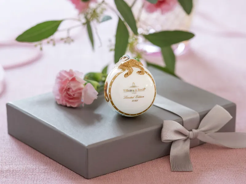 Die Villeroy & Boch Annual Easter Edition zeigt ein dekoratives Ornament mit goldenen Akzenten auf einer grauen Geschenkbox mit Schleife, umgeben von rosa Blumen und grünen Blättern.