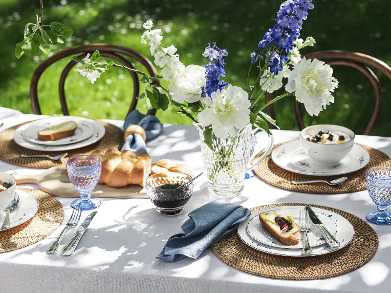 Outdoor table setting with flowers, blue glasses, bread, and Vieux Luxembourg plates with food on woven placemats in a garden setting.