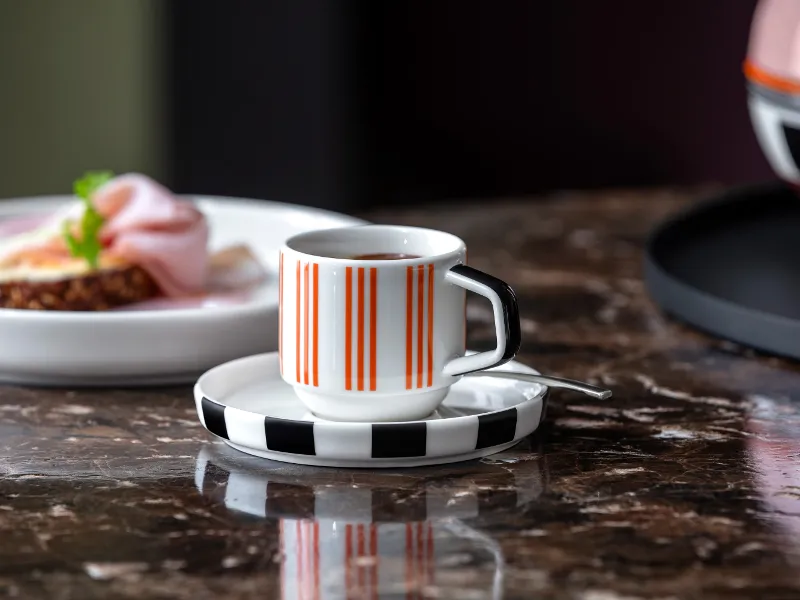 A striped Memphis espresso cup on a saucer sits on a marble table, next to a Memphis plate.

