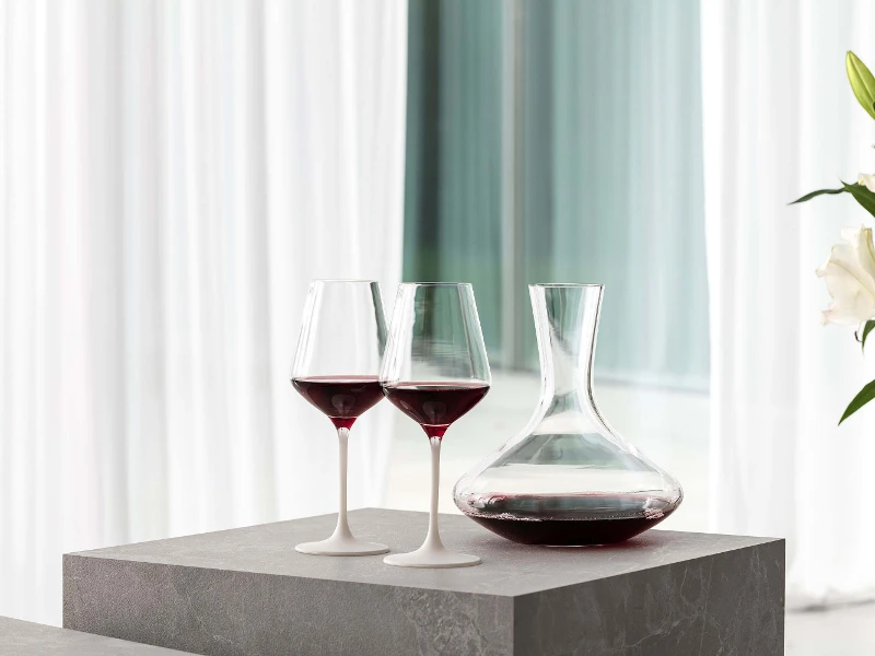 Deux verres à vin rouge Manufacture remplis sur une table posés à côté d’une élégante carafe à vin Purismo.