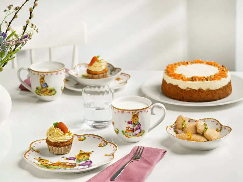 Une table dressée avec des tasses à thé et des assiettes de la collection Annual Easter Edition 2024, avec différentes pâtisseries et une génoise, le tout orné de décorations printanières.