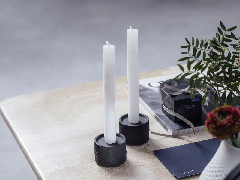 Two white candles in Manufacture candleholders on a marble table, accompanied by a plant.