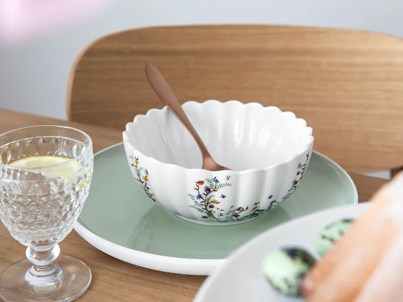 Una ciotola decorativa con motivo floreale della collezione Spring Fantasy di Villeroy & Boch e un cucchiaio su un piatto verde. Un bicchiere d'acqua con limone e un piatto di macarons assortiti si trovano accanto sul tavolo.