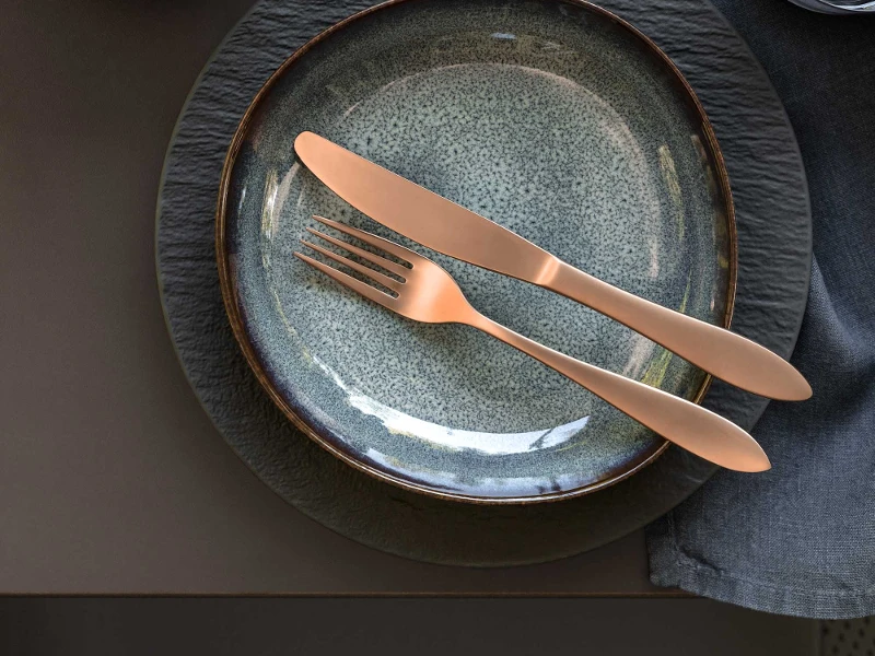 Une décoration de table élégante avec de la vaisselle moderne Manufacture et Lave ainsi que des couverts de table en cuivre de la collection Manufacture sur une nappe foncée.